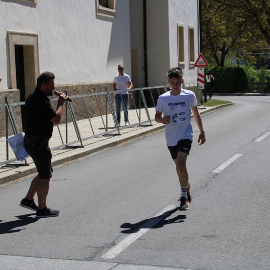 Ein Marathonläufer mit der Nummer 150 auf seinem Shirt joggt eine Straße entlang, während ein Mann mit Mikrofon in der Nähe spricht. Dahinter ist ein Gebäude mit Fenstern und einem Metallzaun zu sehen.