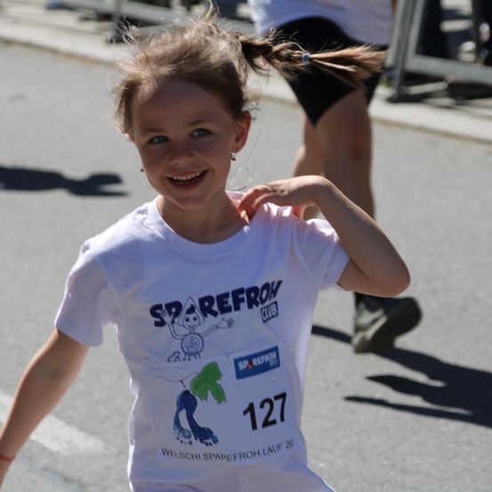 Ein kleines Mädchen mit Zöpfen läuft in einem Rennen, trägt ein weißes T-Shirt mit der Nummer 127 und dem Logo des Sparefroh Clubs.