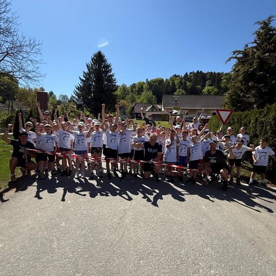 Eine Gruppe von Menschen in passenden weißen T-Shirts und Shorts steht in einer Reihe, einige heben ihre Hände. Sie scheinen an einem sonnigen Tag für ein Foto zu posieren.
