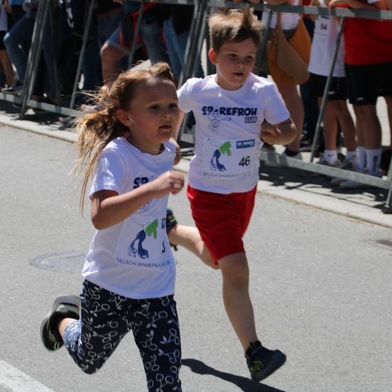 Ein junger Junge und ein Mädchen laufen in einem Marathon und tragen weiße T-Shirts. Dahinter sehen Zuschauer von einer Metallbarriere aus zu.