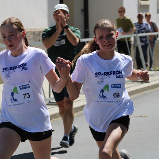 Zwei junge Mädchen laufen einen Marathon und tragen weiße T-Shirts mit den Nummern 73 und 69. Sie halten Händchen, während ein Mann hinter ihnen zusieht.