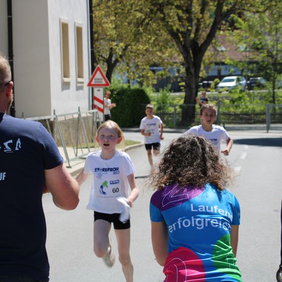 Ein Junge in einem weißen Shirt mit der Nummer 60 läuft in einem Rennen. Ein Mädchen in einem blauen Shirt mit Lauferfolg schaut zu. Ein Mann in einem blauen Shirt geht in der Nähe. Hinter ihnen steht ein Baum und ein Haus.