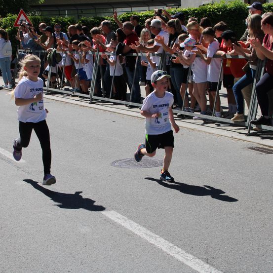 Zwei Kinder rennen bei einem Rennen auf der Straße. Zuschauer beobachten sie von einer Absperrung aus und feuern sie an.