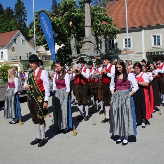 Ein Marschkapelle in traditioneller Kleidung steht auf einem Platz, mit einem Banner und einem Denkmal im Hintergrund. Die Gruppe spielt Musikinstrumente.