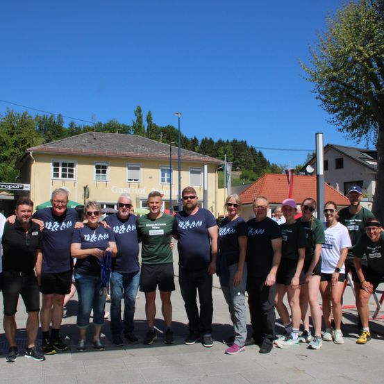 Eine Gruppe von Menschen posiert für ein Foto vor einem Gebäude mit einem roten Dach und einem Baum davor. Sie tragen blaue Shirts mit weißen Buchstaben und einige tragen Mützen.