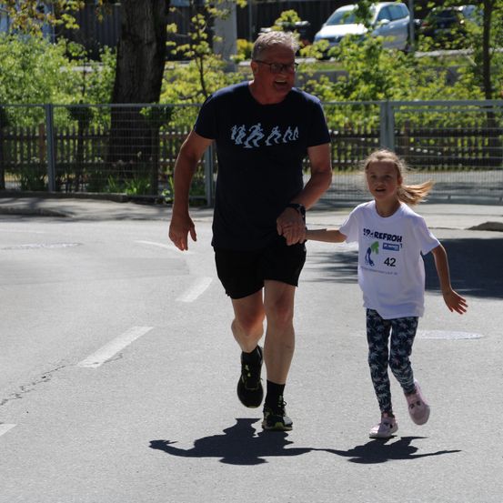 Ein Mann in einem schwarzen T-Shirt läuft mit einem kleinen Mädchen in einem weißen T-Shirt. Sie gehen auf einer gepflasterten Straße, mit Bäumen und geparkten Autos im Hintergrund.