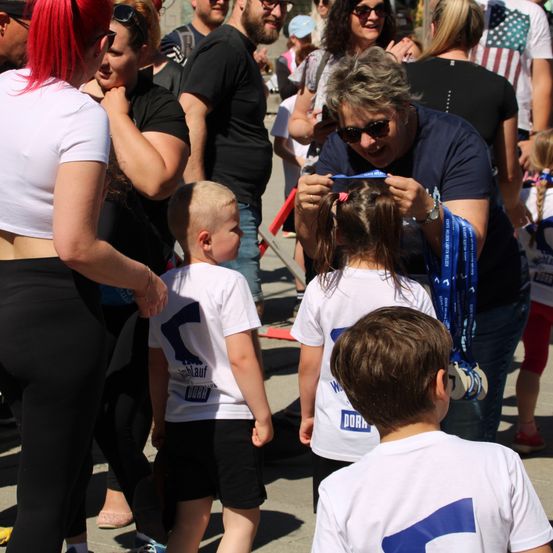 Eine Gruppe von Menschen, einschließlich Kinder in weißen T-Shirts mit dem Wort 'DORA', stehen in einer Reihe. Eine ältere Frau legt ein blaues Band um den Hals eines Kindes.