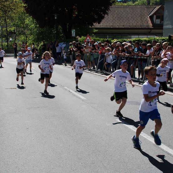 Kinder nehmen an einem Rennen auf einer Straße teil. Zuschauer beobachten von der Seite. Die Kinder tragen weiße Shirts mit Nummern.