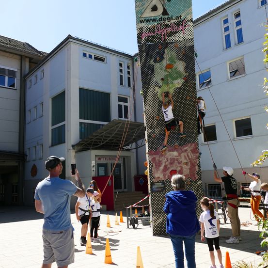 Zwei Personen klettern eine hohe Kletterwand vor einem Schulgebäude. Zuschauer schauen zu und ein Mann macht ein Foto.