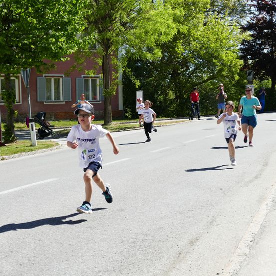 Einige Läufer nehmen an einem Rennen teil. Ein Junge mit der Nummer 122 läuft vorne. Einige Leute stehen am Straßenrand. Eine Frau schiebt einen Kinderwagen.
