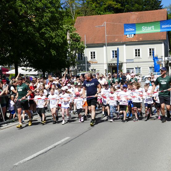 Eine Gruppe von Kindern und Erwachsenen läuft auf der Straße in einem Rennen. Sie passieren ein rotes und weißes Band. Zuschauer beobachten sie von der Seite. Ein Gebäude mit einer Statue ist im Hintergrund.