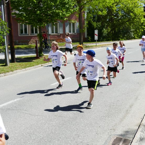 Eine Gruppe von Kindern läuft in einem Rennen auf einer Straße mit Bäumen und einem Gebäude im Hintergrund. Sie tragen alle weiße Trikots mit Nummern.