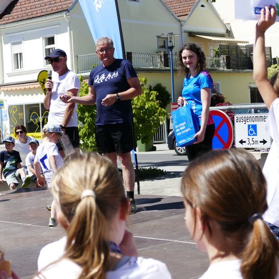 Zwei Erwachsene, einer mit einem Mikrofon, stehen auf einer Bühne und sprechen eine Gruppe von Kindern an, die in der Nähe sitzen und stehen. Eine Frau hält eine blaue Tasche mit einem Logo. Dahinter sind Gebäude und ein blauer Banner zu sehen.