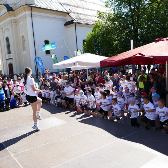 Eine Frau in Weiß und Schwarz führt eine Gruppe von Kindern vor einem Gebäude unter Schirmen. Die Kinder tragen weiße Hemden und Shorts.