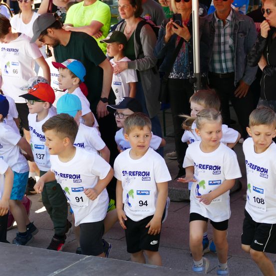 Eine Gruppe von Kindern, die weiße T-Shirts mit der Nummer und dem Text 'Sp.Refroh Club' tragen, steht in einer Schlange. Dahinter stehen mehrere Erwachsene, einige mit Mützen und Sonnenbrillen. Die Kinder schauen in verschiedene Richtungen.