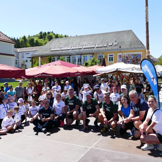 Gruppenfoto von Läufern und Unterstützern an einem sonnigen Tag. Rote und weiße Regenschirme sind zu sehen. Ein Gebäude mit Solarmodulen im Hintergrund.