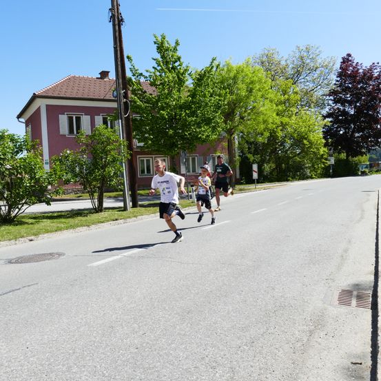 Drei Personen laufen auf der Straße. Ein Junge in der Mitte trägt einen Hut. Hinter ihnen ist ein Haus mit roten Wänden und weißen Fensterläden zu sehen. Bäume säumen die Straße.