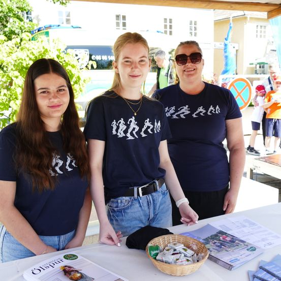Drei Frauen stehen lächelnd hinter einem Tisch. Sie tragen marineblaue T-Shirts mit Lauffiguren. Ein Korb mit Gegenständen und Broschüren steht auf dem Tisch. Im Hintergrund sind ein Lieferwagen und ein Gebäude mit Fahnen zu sehen.