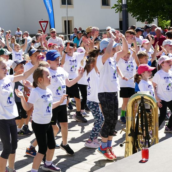 Eine Gruppe von Kindern und Erwachsenen in weißen T-Shirts tanzt bei einer Parade. Einige von ihnen heben die Hände, während andere Mützen tragen. Eine Person vorne spielt Tuba. Der Boden ist mit Beton gepflastert.