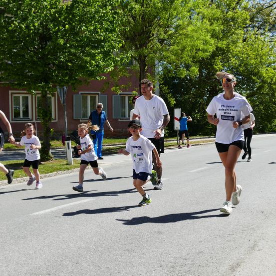 Ein Marathon mit Läufern auf einer Stadtstraße. Ein Mann und eine Frau in der Führung tragen Sonnenbrillen und lächeln. Dahinter sind Kinder und Erwachsene, darunter ein Mann in einer blauen Weste.