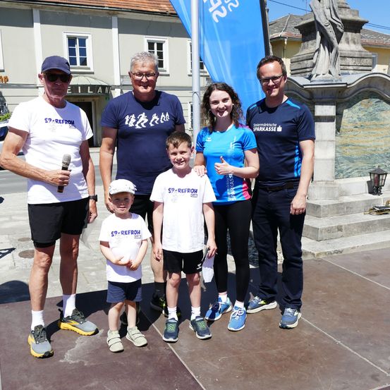 Fünf Personen, darunter zwei Kinder, posieren für ein Foto auf einem Bürgersteig. Dahinter eine Statue, eine blaue Fahne und ein Gebäude.