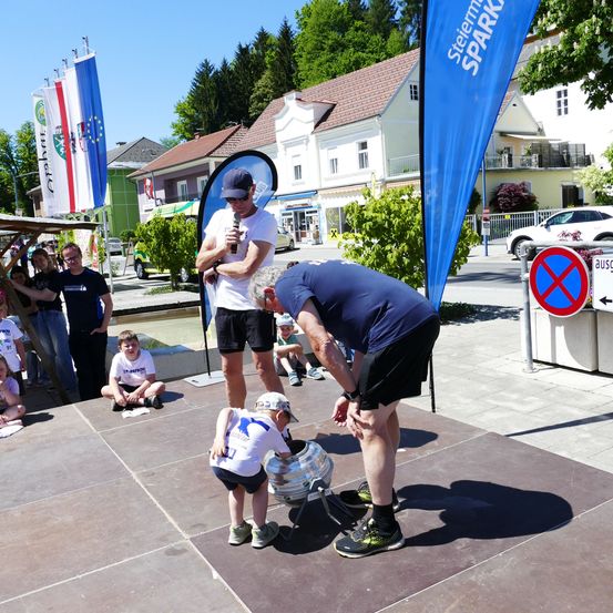 Ein junger Junge wird von einem Mann in Blau unterstützt, der ihm beim Heben eines großen Fasses hilft. Im Hintergrund beobachtet eine Menge.