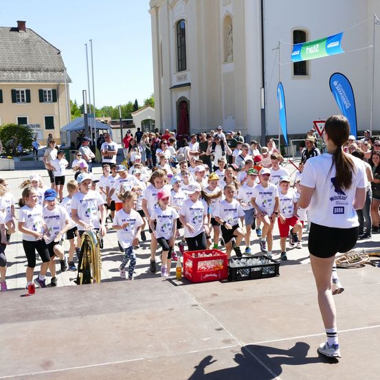 Eine Gruppe von Kindern in weißen Shirts und Mützen läuft vor einer Menge und Gebäuden in einem Rennen. Eine Frau in Weiß geht hinter ihnen.