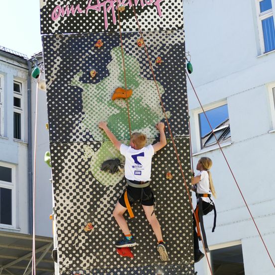 Zwei Kinder klettern an einer farbenfrohen Wand mit grünen Mustern und tragen Sicherheitsgurte vor einem Gebäude.