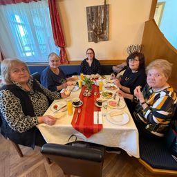 Fünf Frauen sitzen an einem Tisch und genießen ihre Mahlzeit mit verschiedenen Speisen und Getränken. Eine Frau hält ein Glas Orangensaft.