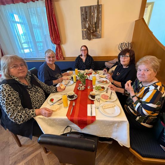 Fünf Frauen sitzen an einem Tisch und genießen ihre Mahlzeit mit verschiedenen Speisen und Getränken. Eine Frau hält ein Glas Orangensaft.