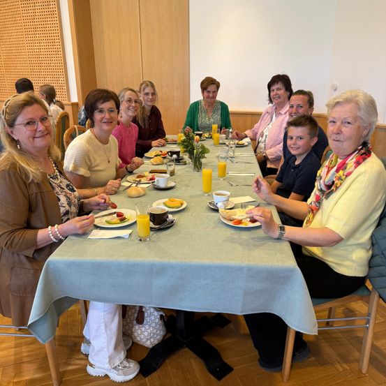 Mehrere Frauen und ein Junge sitzen an einem Tisch, essen und trinken. Sie haben Teller mit Essen, Gläser Orangensaft und eine Vase mit Blumen auf dem Tisch. Der Raum ist hell mit hölzernen Wänden.