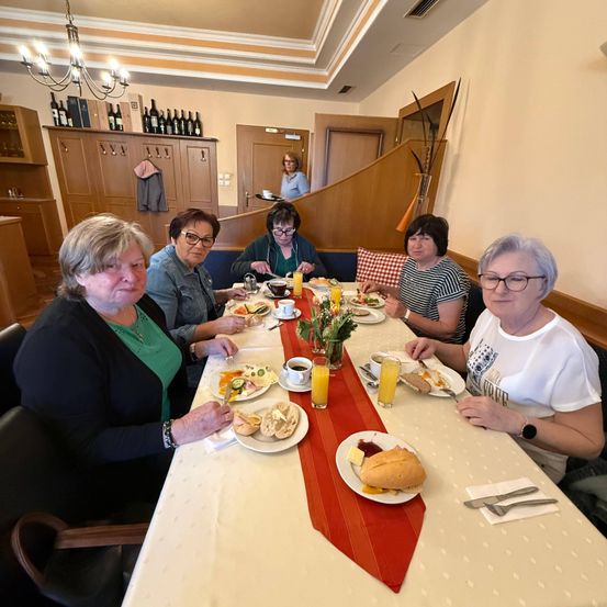 Eine Gruppe von Frauen sitzt an einem langen Tisch in einem Essbereich und isst. Es gibt Teller mit Essen und Getränken auf dem Tisch. Die Frau auf der linken Seite hält Messer und Gabel.