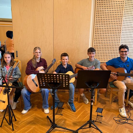 Fünf Personen, wahrscheinlich Schüler, sitzen gemeinsam auf Stühlen mit Gitarren. Sie haben Notenständer vor sich und scheinen sich in einem Studio zu befinden.