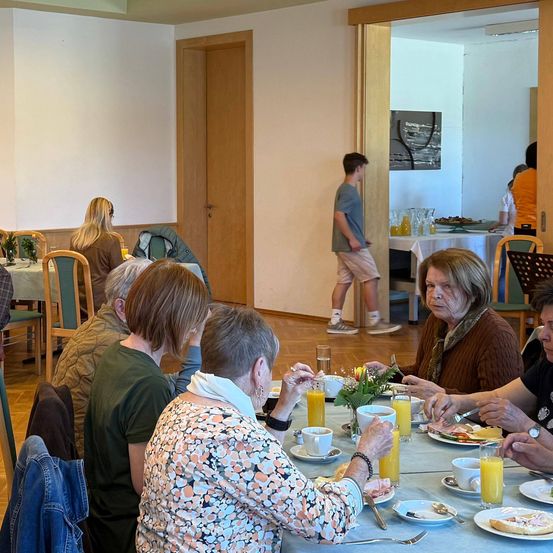 Eine Gruppe von Menschen sitzt um einen Esstisch herum, isst und trinkt. Eine Person steht in der Nähe einer Tür, eine andere an einem Tisch mit Blumenarrangement.