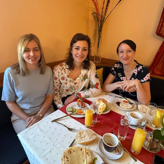 Drei Frauen sitzen lächelnd an einem Tisch. Der Tisch hat Teller mit Essen, Tassen und Gläser. Eine Frau hält einen Löffel und der Tisch ist mit einem roten Tuch gedeckt.