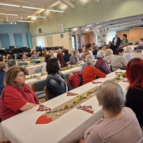 Ein Indoor-Event mit einem langen Tisch für viele Gäste, eine Gruppe von Menschen sitzt, und einige stehen im Hintergrund. Der Tisch hat eine weiße Tischdecke, Blumen und Besteck.