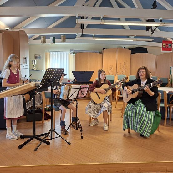 Vier Personen in einem Raum spielen verschiedene Musikinstrumente. Eine Frau spielt Gitarre, eine andere Akkordeon und ein Mädchen Zither. Ein Pokal und Bücher sind ebenfalls vorhanden.