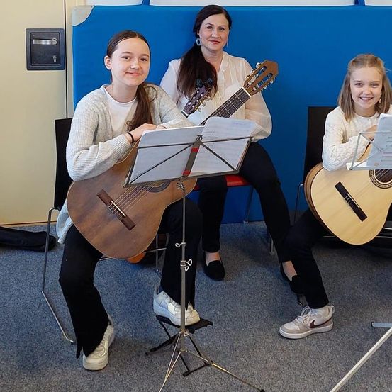 Drei Personen sitzen und lächeln, halten Gitarren. Die Frau in der Mitte hat eine Gitarre auf dem Schoß. Sie befinden sich wahrscheinlich in einem Musikraum mit Notenständern vor ihnen.