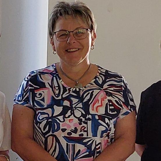 Drei Frauen in Schürzen stehen zusammen drinnen. Die Frau auf der linken Seite trägt ein weißes Hemd, die in der Mitte ein farbenfrohes Hemd, und die auf der rechten Seite ein schwarzes Hemd. Sie stehen neben einem Tisch mit Tellern.