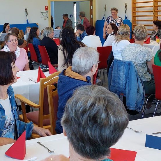Eine Gruppe von Frauen in Freizeitkleidung sitzt in einem Esszimmer an Tischen mit weißen Tischdecken, roten Servietten und Besteck. Einige unterhalten sich, während andere sich auf ihr Essen konzentrieren. Der Raum hat Holzstühle, eine Holztür und verschiedene Gegenstände an den Wänden.