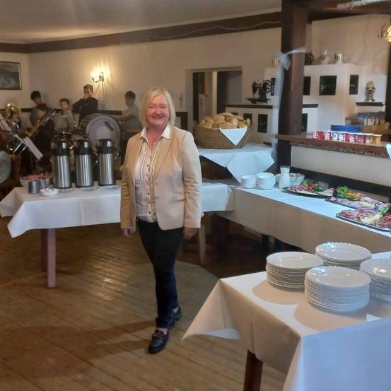 Eine lächelnde Frau steht in einem Raum in der Nähe eines Buffet-Tisches, mit einer Band im Hintergrund. Der Tisch hat Teller, Schüsseln und einen Korb mit Brot.