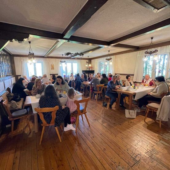 Ein Restaurant mit Holzboden, Tischen mit weißen Tischdecken und Kronleuchtern. Die Leute sitzen und scheinen eine Mahlzeit zu sich zu nehmen. Der Raum ist gut beleuchtet und hat eine rustikale Atmosphäre.