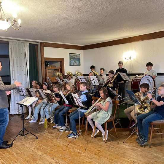 Eine Gruppe von Kindern spielt verschiedene Musikinstrumente in einem Raum mit Holzböden. Ein Mann steht und scheint sie zu dirigieren.