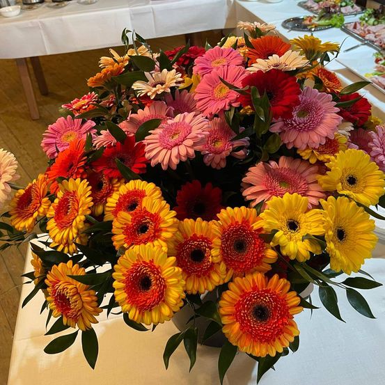 Eine farbenfrohe Blumenarrangement aus mehrfarbigen Gänseblümchen auf einem Tisch, mit grünen Blättern und einer weißen Tischdecke, in einem Raum mit Holzböden.
