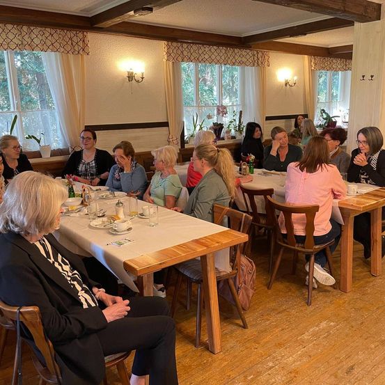 Eine Gruppe von Frauen sitzt um einen Tisch in einem Restaurant. Sie sitzen an langen Holztischen mit weißen Tischdecken. Sie unterhalten sich und lachen. Der Raum hat Holzböden, Holzbalken und weiße Wände.