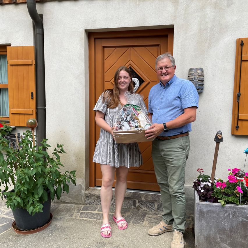 Ein Mann und eine Frau stehen lächelnd vor einem Haus. Die Frau hält einen Korb mit einem Geschenk. Der Mann trägt eine Brille und eine Uhr. Dahinter stehen Pflanzen und eine braune Tür.