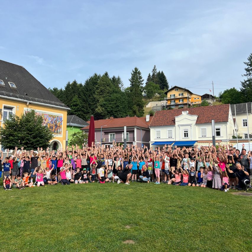 Eine Gruppe von Menschen posiert für ein Foto auf einem Rasenfeld mit Gebäuden im Hintergrund unter einem bewölkten Himmel.