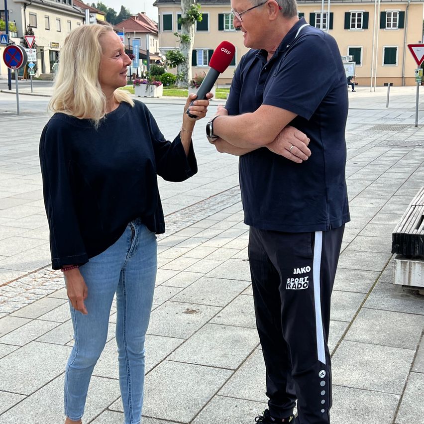Eine Frau mit blonden Haaren führt ein Interview mit einem Mann, der ein blaues Polohemd und schwarze Hosen trägt, auf einer gepflasterten Straße. Hinter ihnen befindet sich ein Gebäude mit mehreren Fenstern.