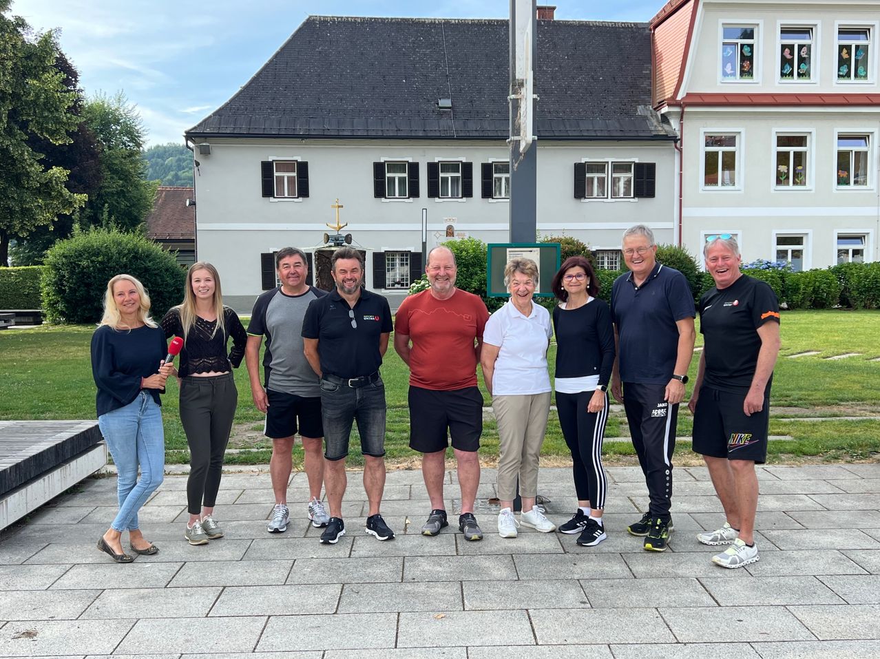 Eine Gruppe von Menschen, möglicherweise eine Familie, steht vor einem Haus. Eine Person hält ein Mikrofon. Sie alle lächeln für ein Foto.