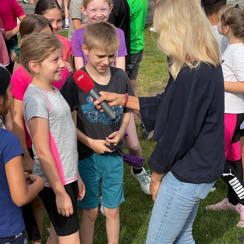 Eine Frau mit Mikrofon interviewt einen Jungen und ein Mädchen draußen, umgeben von Kindern auf einem Rasen.